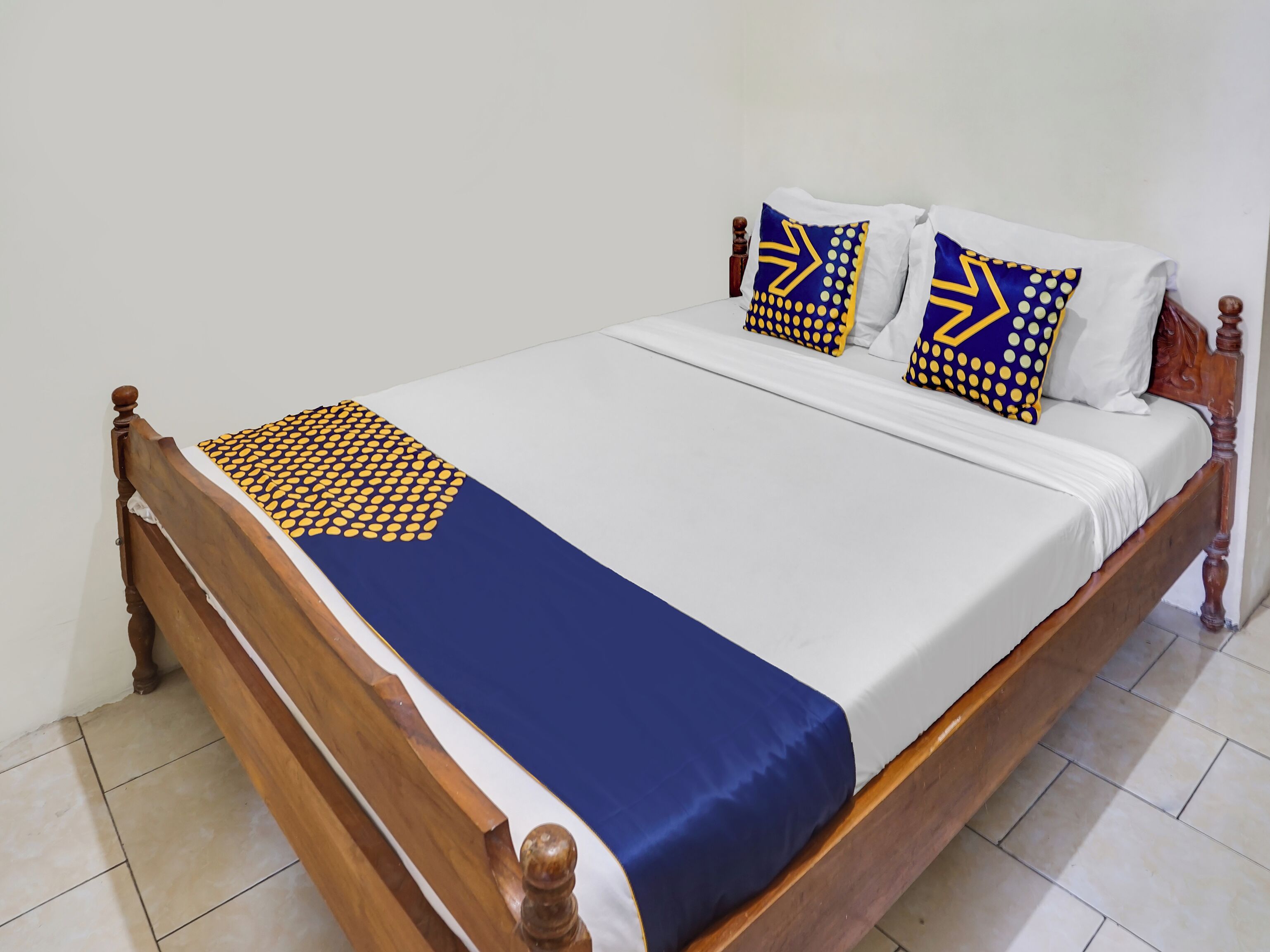 Ein Bett mit Holzbettgestell, weißer Bettwäsche und Kissen mit blau-gelbem Muster.