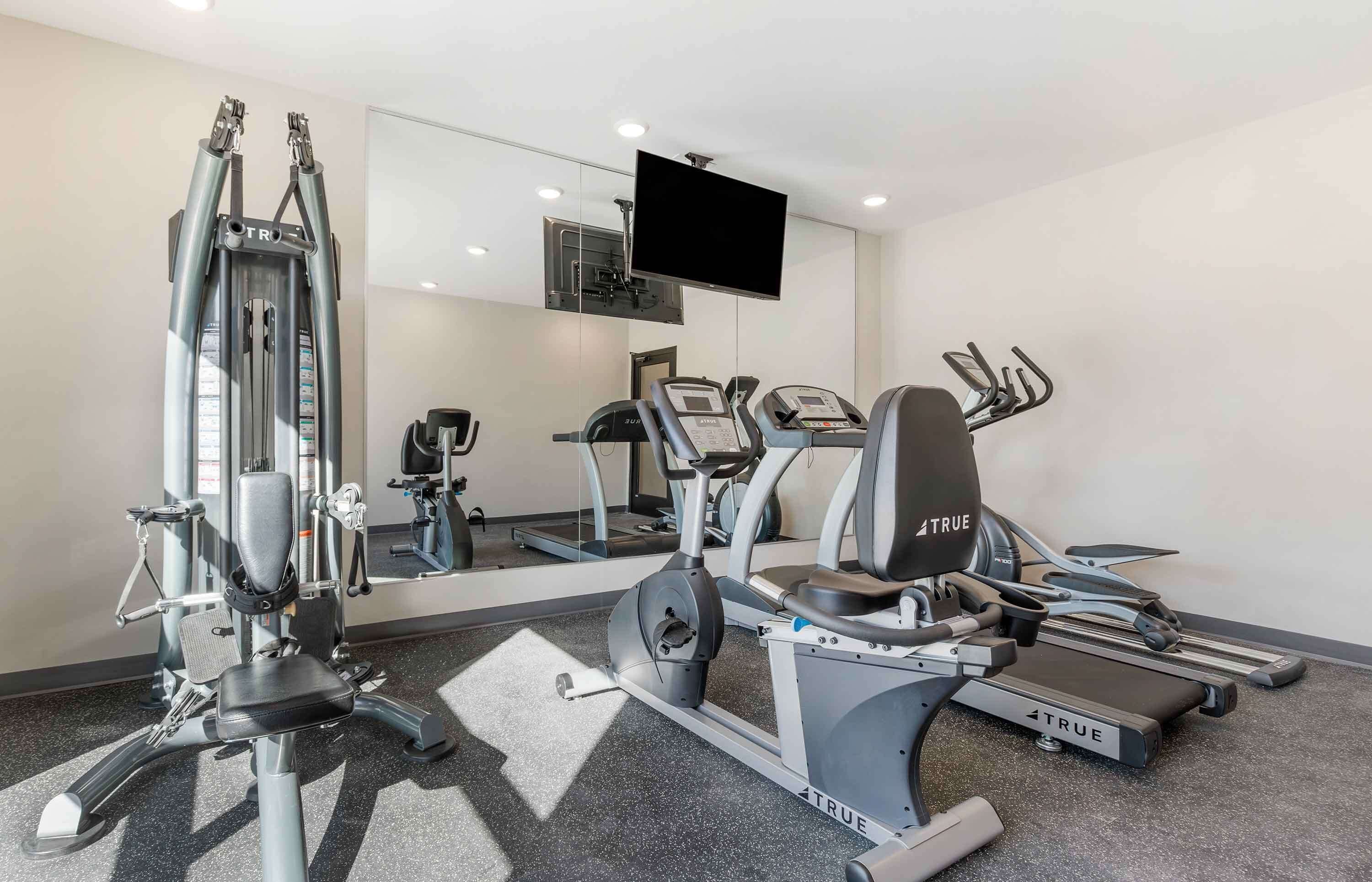 Un gimnasio con diversas máquinas de ejercicio, un espejo grande y un televisor montado.