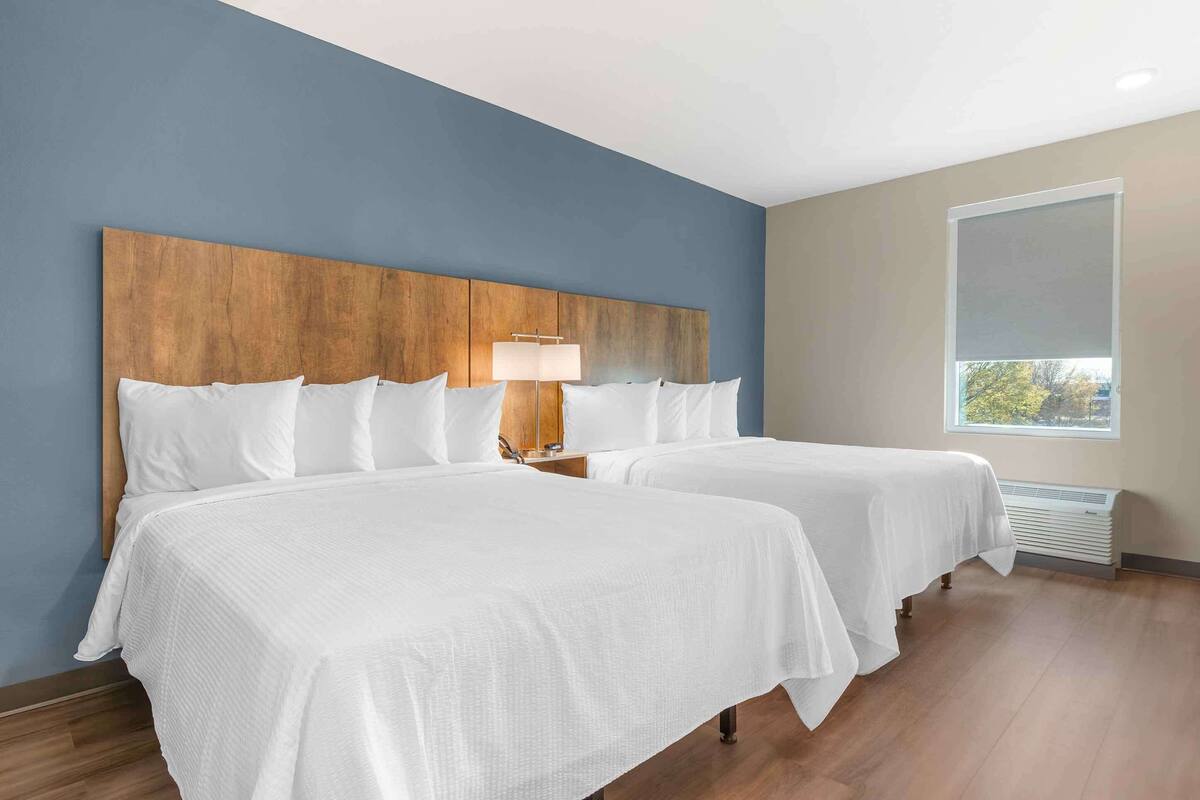 Habitación de hotel con dos camas, cabecero de madera, mesita de noche con lámpara, ventana con vistas y aire acondicionado integrado.