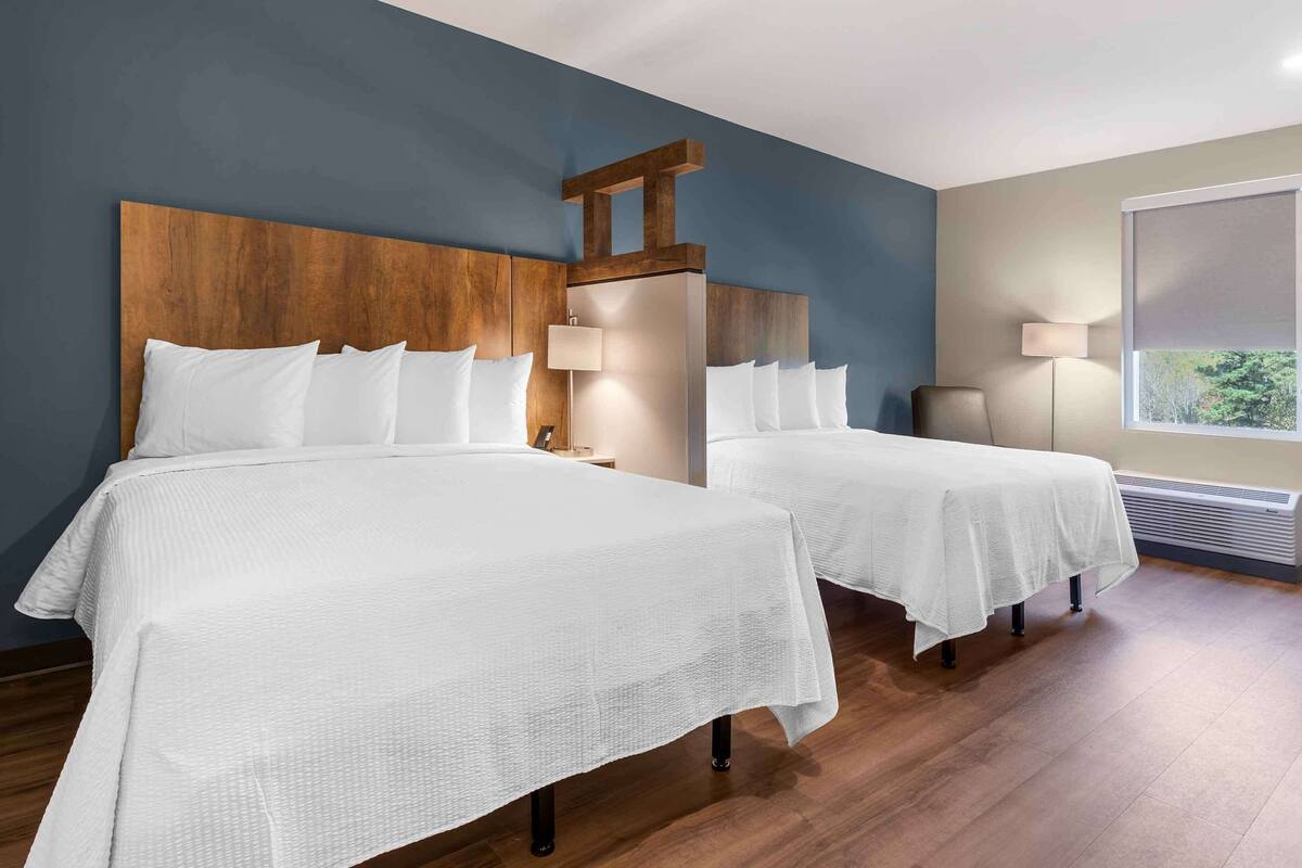 Habitación de hotel con dos camas, cabecero de madera, mesita de noche, lámpara y ventana con vistas.