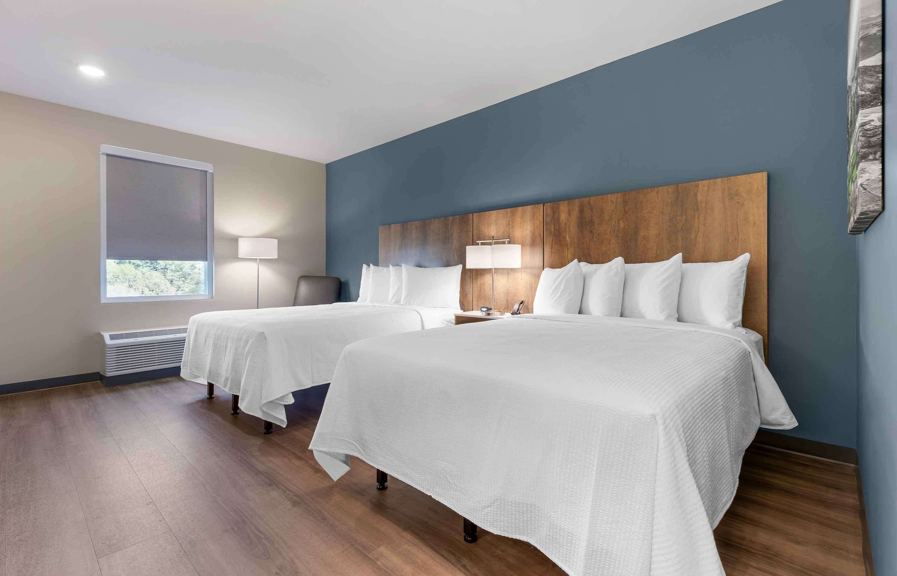 Habitación de hotel con dos camas, un ventanal con persianas, una lámpara de pie y una obra de arte en la pared.