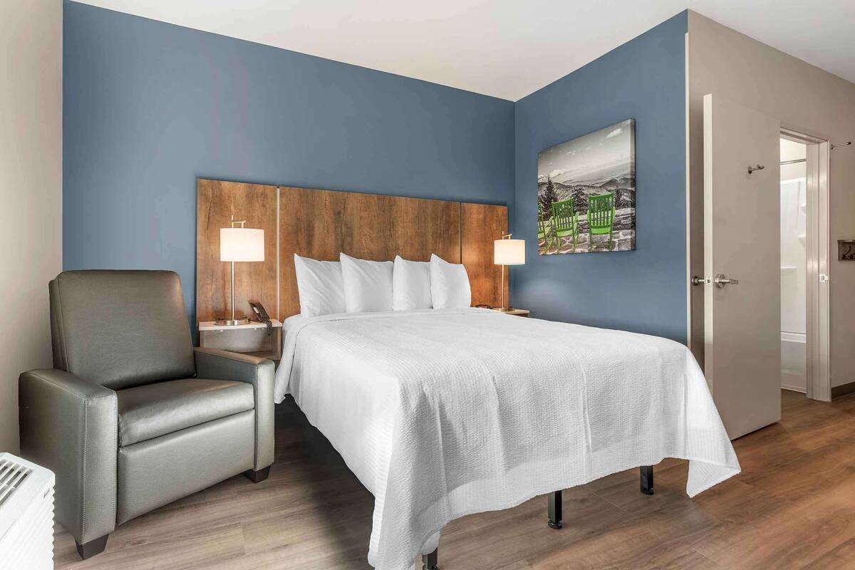 Una habitación de hotel con cama, una silla, una mesita de noche, una lámpara y un cuadro en la pared.
