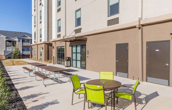 A modern outdoor patio area with picnic tables and chairs, a round table, and a building with multiple floors and balconies.