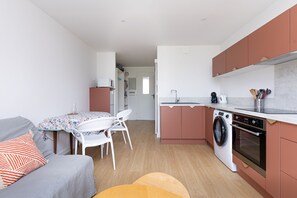 A compact kitchen with a washing machine, a dining area with a table and chairs, and a grey sofa with an orange pillow. - Le Balcon du Port - Joli studio à Capbreton (Capbreton)