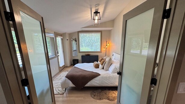 A bedroom with a bed, bedside tables, a nightstand, a window with blinds, and a ceiling light fixture.