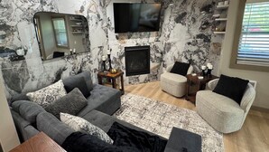 A living room with a grey sofa, a white armchair, a flat-screen TV mounted on the wall, and a fireplace.
