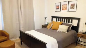 A bedroom with a bed, a nightstand, a chair, and a wall with three framed pictures.