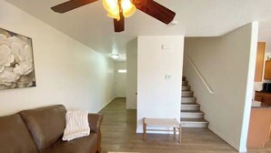 A living room with a brown leather sofa, a wooden coffee table, and a ceiling fan. A staircase leads to a kitchen area.