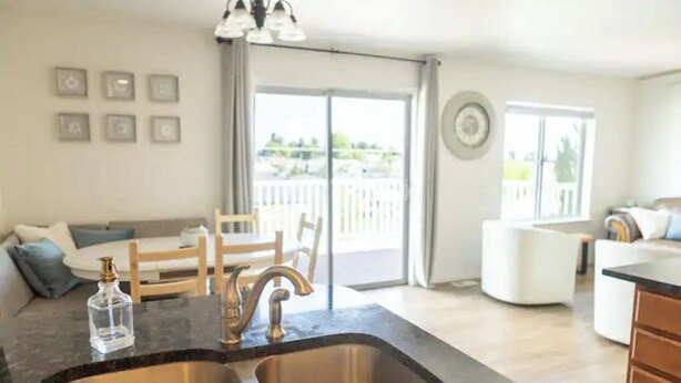 A modern kitchen with a double sink, a dining area with a table and chairs, and a view of a balcony through large sliding doors.