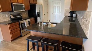 A kitchen with wooden cabinets, a stainless steel microwave, a refrigerator, a sink, and three bar stools.