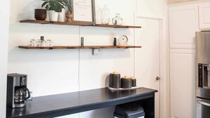 A kitchen with white cabinetry, wooden shelves, a black countertop, a coffee maker, a trash bin, and a stainless steel refrigerator.