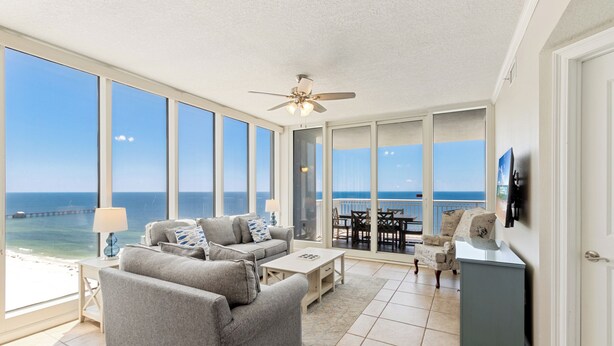 A spacious living room with a large window overlooking the ocean, a ceiling fan, and a dining area with outdoor seating. - The Colonnades 1901 (Gulf Shores)