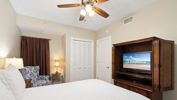 A hotel room with a large bed, a ceiling fan, a television, a desk with a lamp, patterned chairs, and a door with louvered shutters. - The Colonnades 1901 (Gulf Shores)
