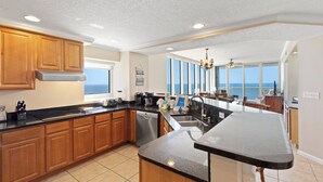 A modern kitchen with wooden cabinets, a black countertop, and a large island. There is a view of the ocean through large windows. - The Colonnades 1901 (Gulf Shores)