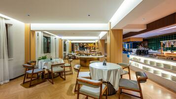 Un comedor moderno con mesas redondas y sillas de madera, una cocina al fondo y un mostrador tipo buffet con platos apilados.