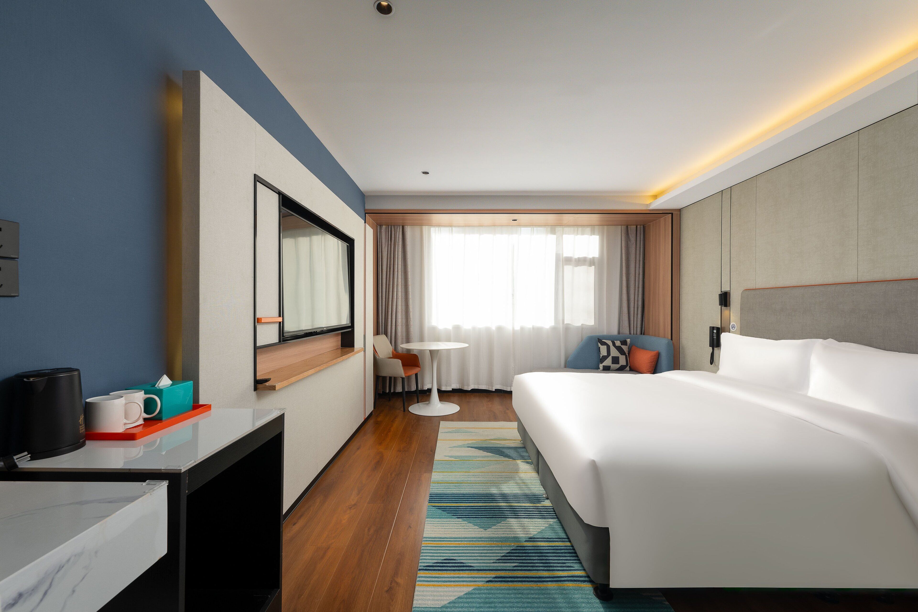 A modern hotel room with a large bed, a desk with a coffee maker, a small table, and a view of the outdoors through the window.