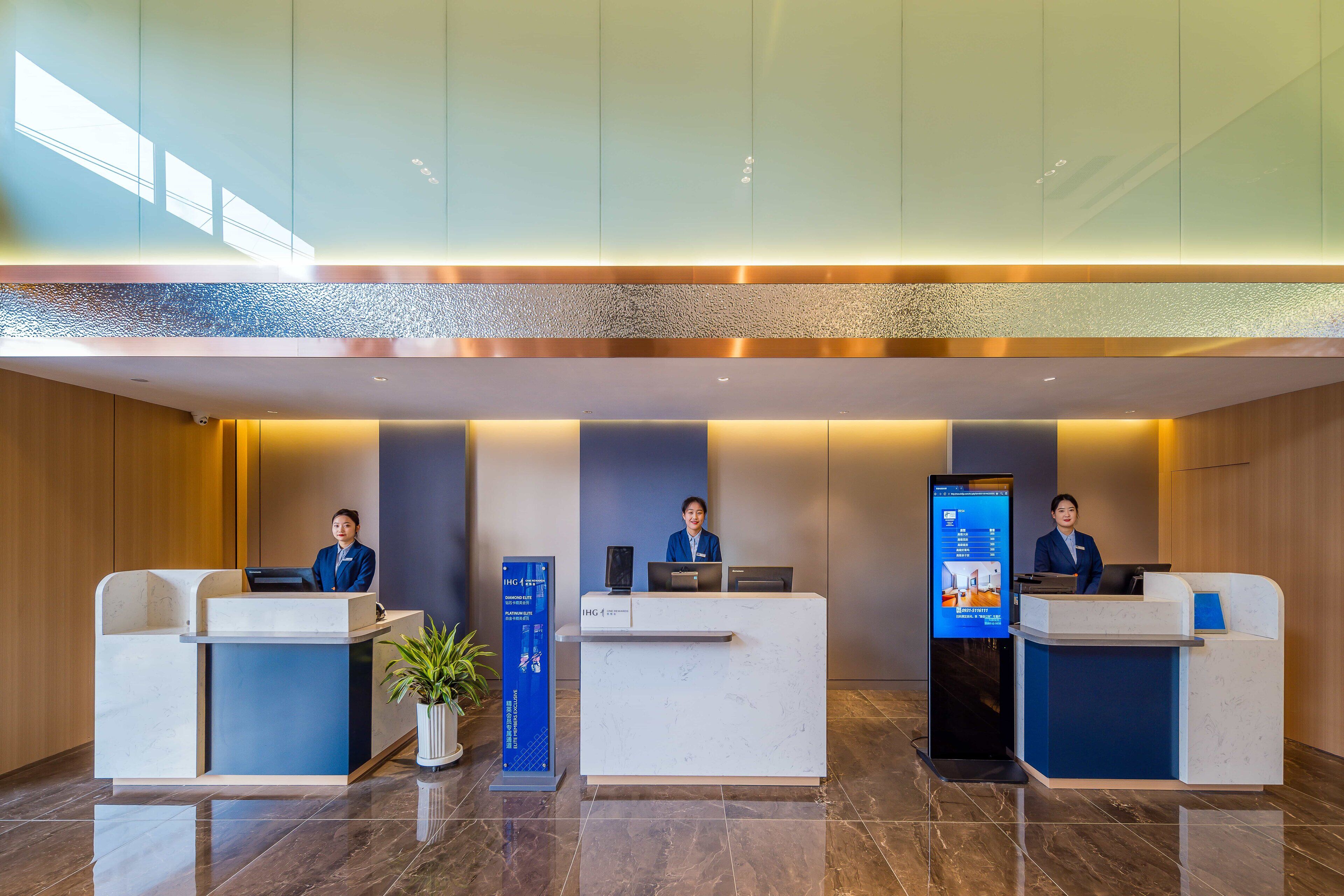 Zona de recepción de un hotel con tres mostradores, cada uno atendido por un recepcionista. A la izquierda, un quiosco de información digital y, a la derecha, una pantalla grande que muestra información del establecimiento.