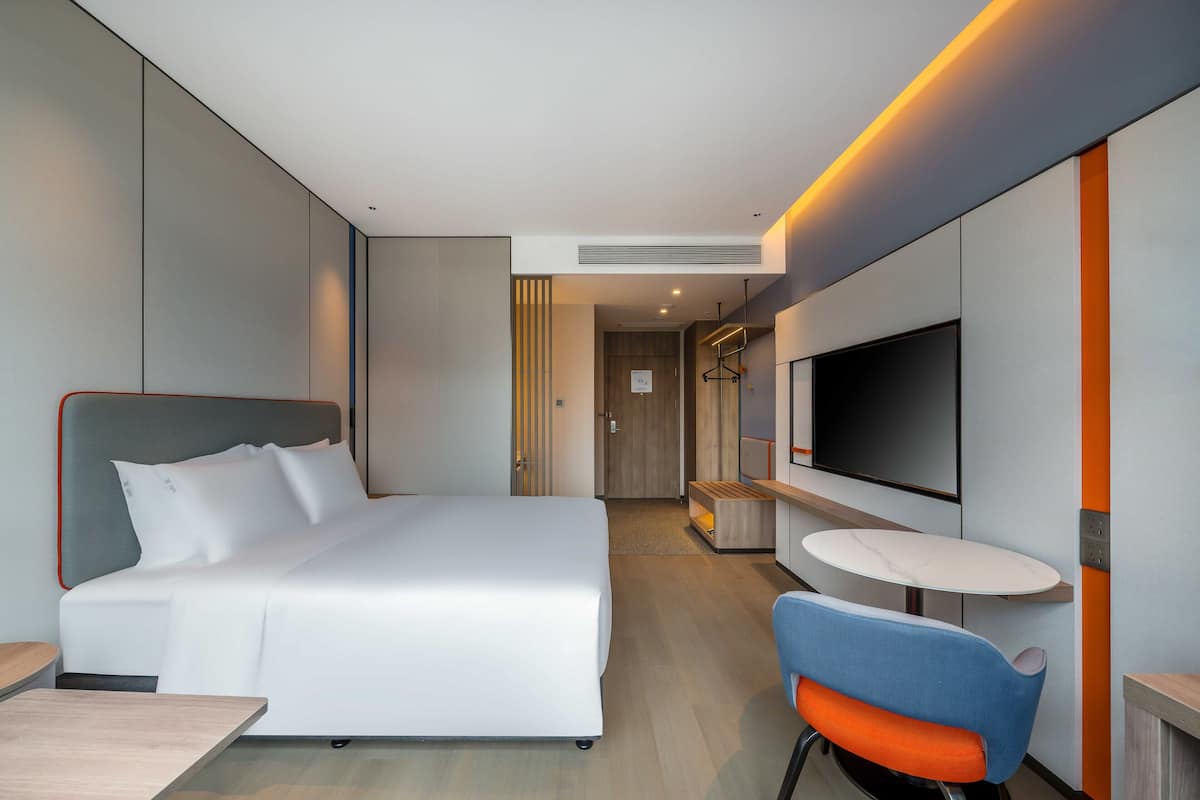 A modern hotel room with a large bed, a TV mounted on the wall, a small round table, and a blue chair.