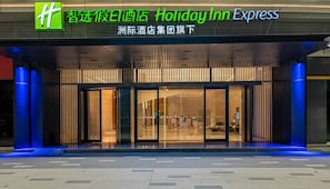 Entrada de un hotel Holiday Inn Express con un diseño moderno, destacando amplias puertas de vidrio y un interior acogedor visible desde el acceso.