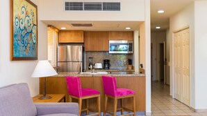 A compact kitchen with wooden cabinets, a stainless steel refrigerator, a microwave, and a coffee maker. There are two pink bar stools and a table with a lamp.