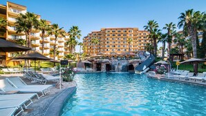 A hotel pool area with lounge chairs, a waterfall feature, and a large building in the background.