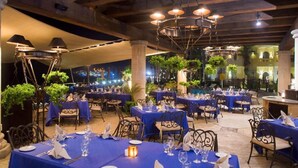 An indoor dining area with tables set for service, wooden beams on the ceiling, and potted plants.