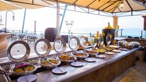 A buffet setup with various food dishes under a covered area with a view of the ocean.