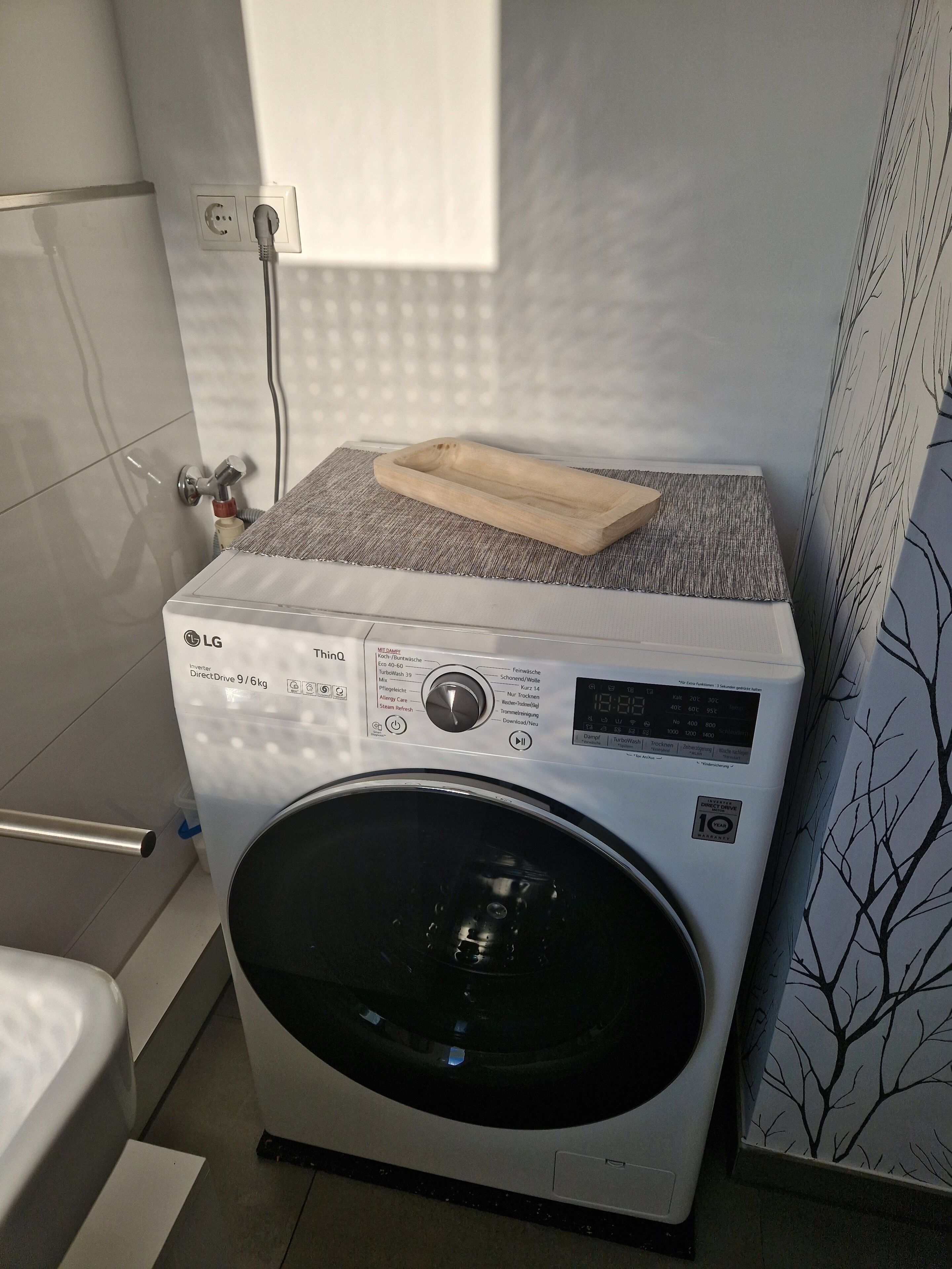 Eine kompakte, weiße LG DirectDrive Waschmaschine mit 9 kg Frontlader und Bedienfeld, platziert in einem kleinen Waschraum mit gefliesten Wänden und einem Fenster.