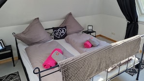 A single bed with a woven headboard, two pillows, a small bedside table with a picture frame, and a window with curtains.