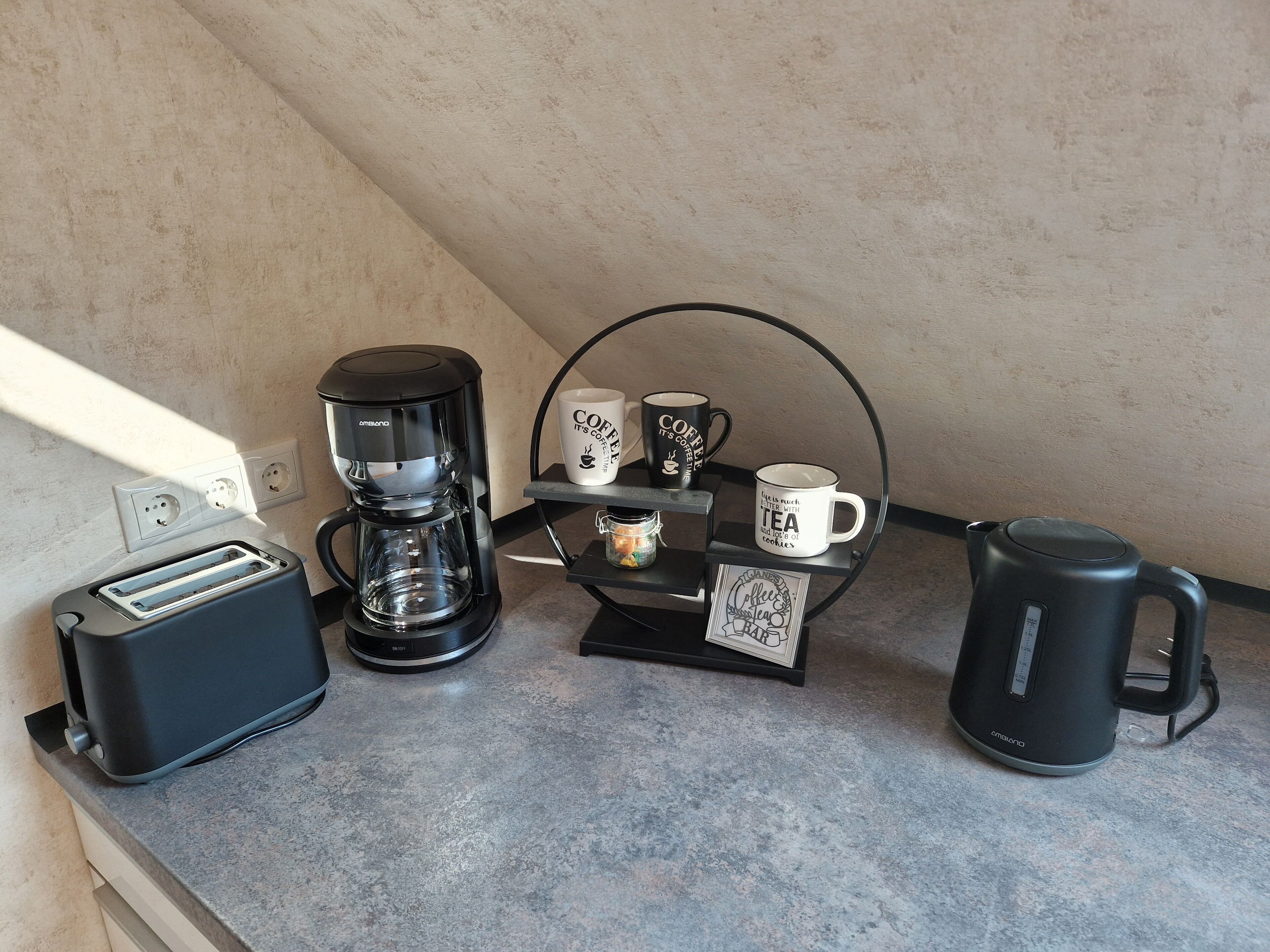 A kitchenette with a coffee maker, a kettle, a toaster, and a mug rack with coffee and tea mugs.