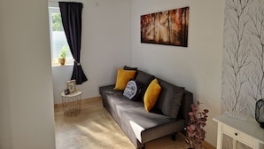 A living room with a grey sofa, yellow and black cushions, a painting of a forest, a small round table with a plant, and a white side table with a fan.