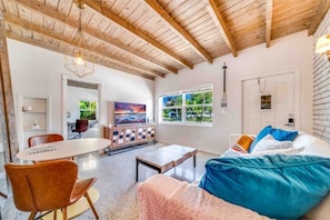 Ein geräumiges Wohnzimmer mit Holzboden, einem großen Fenster mit Blick auf Grünflächen, einem Sofa mit bunten Kissen und einem Fernseher auf einer Holzkommode.