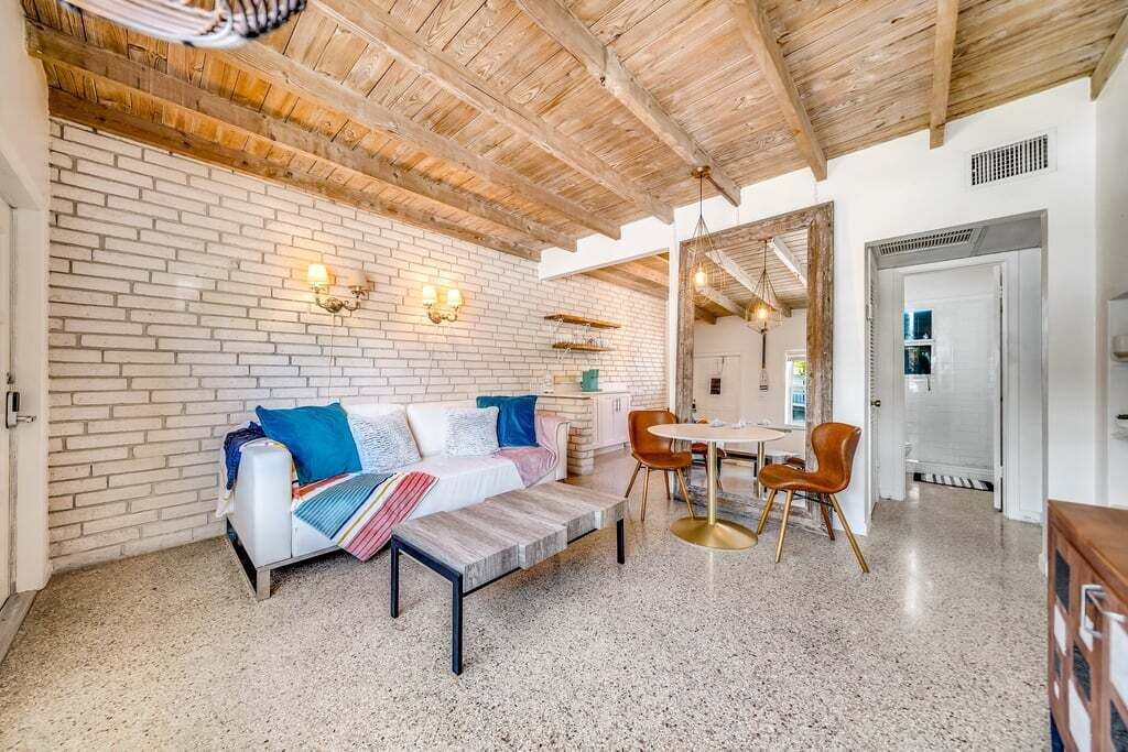 Ein modernes Wohnzimmer mit Backsteinwand, Holzbalkendecke, einem weißen Sofa mit bunten Kissen, einem Couchtisch und einem Essbereich mit Stühlen und Tisch.