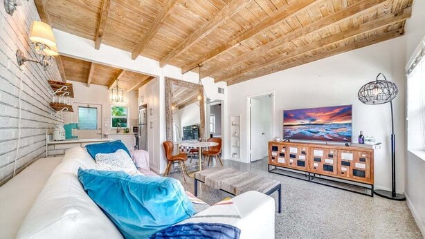 Uma sala de estar moderna com teto de madeira, um sofá branco com almofadas azuis, uma área de jantar com mesa e cadeiras, e uma televisão sobre um armário de madeira.