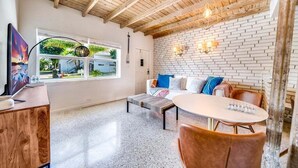 Uma sala de estar moderna com parede de tijolos brancos, vigas de madeira no teto, uma mesa redonda e um sofá com almofadas coloridas.