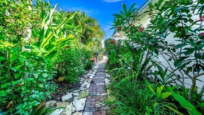 Um caminho de pedra estreito, ladeado por vegetação exuberante e um edifício branco.