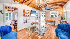 Ein geräumiges Wohnzimmer mit einem Glastisch, blauen Sofas, Holzböden und einer hohen Decke mit freiliegenden Balken.