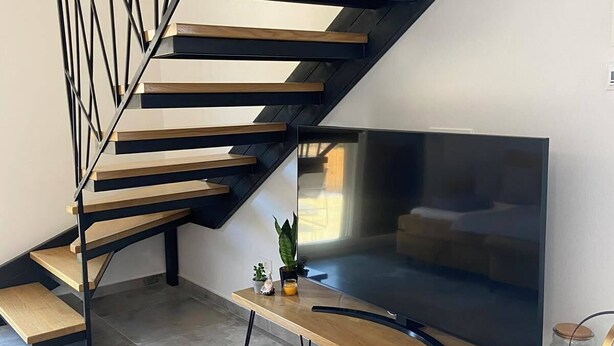 Una moderna scala con gradini in legno e ringhiere in metallo nero, che conduce a una zona living con una grande TV a schermo piatto, un tavolino con una pianta e un telecomando sul pavimento.