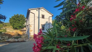 Un piccolo edificio a un piano con facciata bianca e tetto spiovente, immerso nel verde e circondato da un muro in pietra.