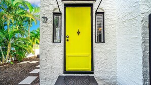 Une porte jaune vif, ornée d’une poignée de porte noire, est encadrée de deux fenêtres aux montures noires et aux volets blancs, le tout intégré dans un mur recouvert d’un enduit blanc.