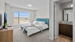 Een slaapkamer met een groot raam, een bed met een gestapeld hoofdeinde, een televisie en een badkamer met een spiegel en wastafel.