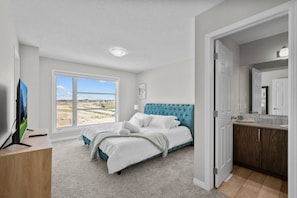 Ein Schlafzimmer mit großem Fenster, einem Bett mit gestepptem Kopfteil, einem Fernseher und einem Badezimmer mit Spiegel und Waschbecken.