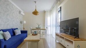 A modern living room with a blue sofa, a wooden coffee table, a flat-screen TV on a wooden stand, and a dining area with a white table and chairs.