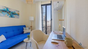 A modern living room with a blue sofa, a wooden desk with a laptop, a small table with a plant, and a large window with a view of buildings.