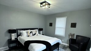Une chambre à coucher avec un grand lit, une table de chevet, une chaise, une fenêtre avec des stores et un tapis décoratif.