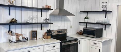 A modern kitchen with white cabinetry, a stainless steel stove, and a microwave. There are wooden shelves with decorative items and a black ceiling light fixture.