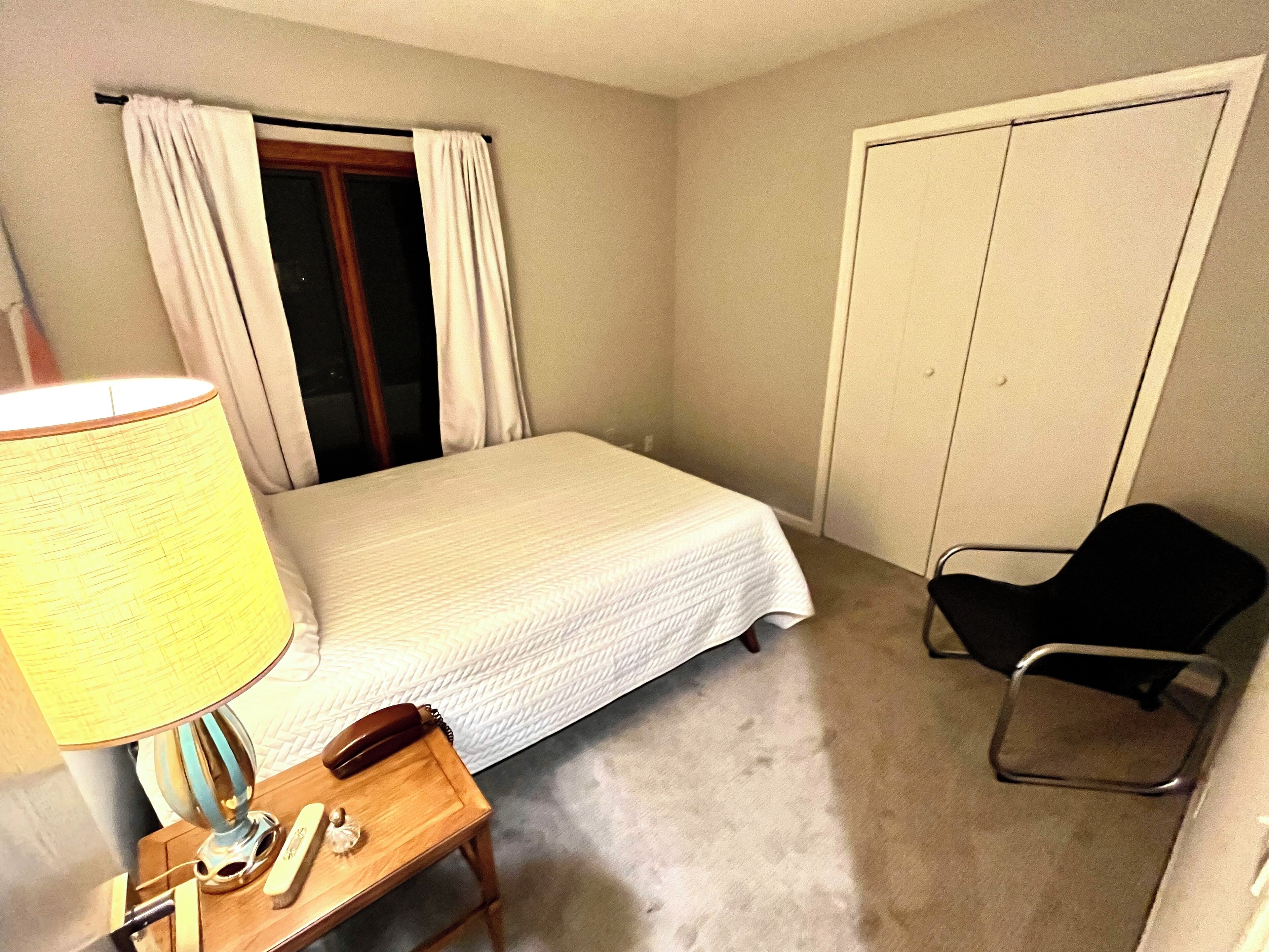 A small, neatly arranged hotel room with a single bed, a chair, a bedside table with a lamp, and a telephone.