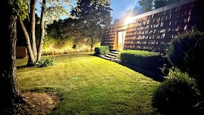 A well-lit garden area with a wooden fence, a grassy lawn, and a pathway leading to a building entrance.