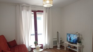 Ein Wohnzimmer mit einem roten Sofa, einem Fernseher auf einem weißen Schrank, einem kleinen Tisch mit einer Pflanze und einem Fenster mit Vorhängen.