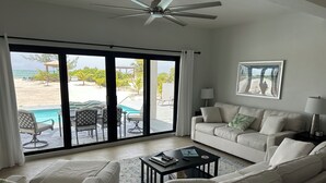A spacious living room with a large sliding glass door leading to a pool area, a ceiling fan, a beige sofa, and a framed picture on the wall. - BRAND NEW Beachfront Villa best sandy beach in TCI with pool Fox Tail Palm Villa (Whitby)